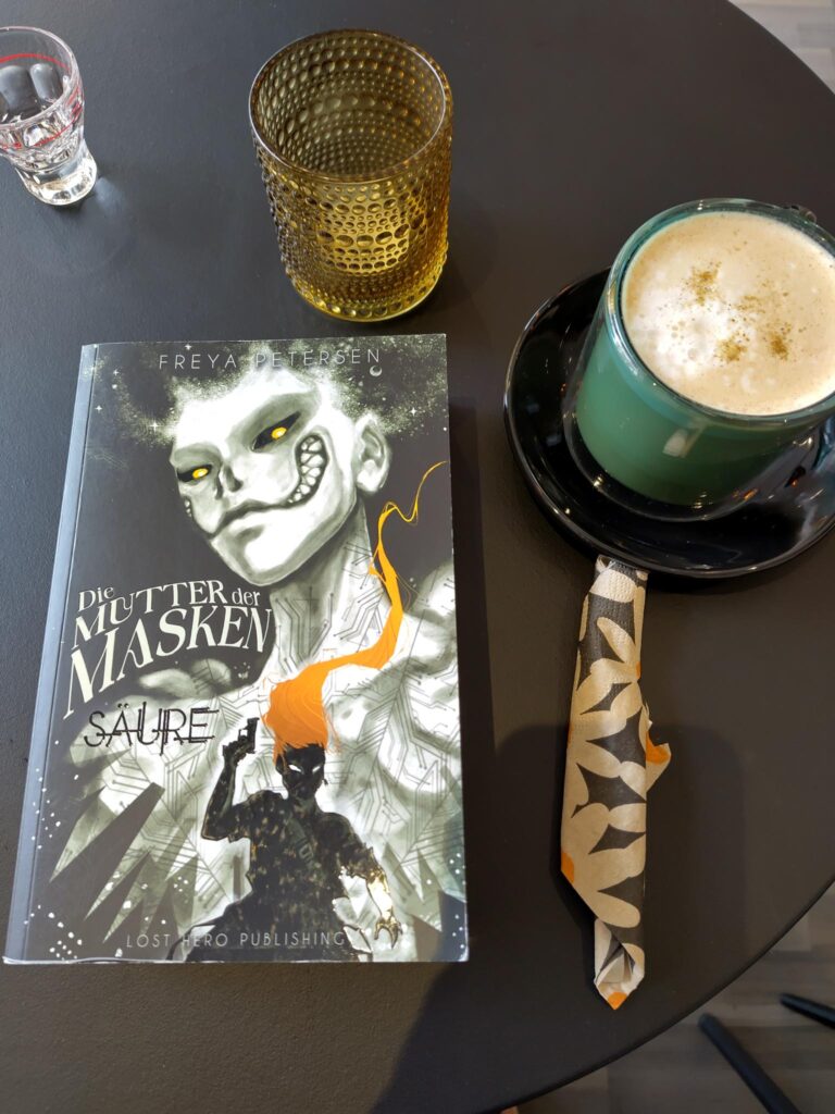 Das Buch "Die Mutter der Masken" liegt auf einem Tisch neben einer Kaffeetasse.