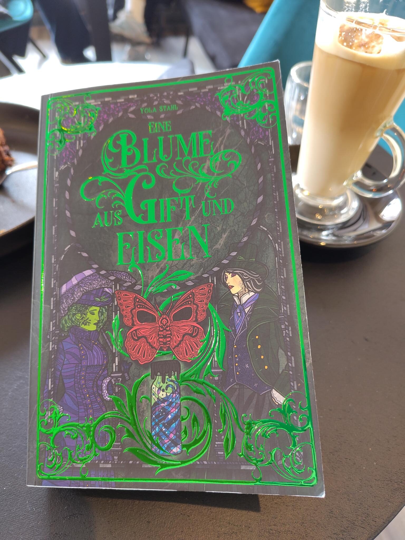 Das Buch "Eine Blume aus Gift und Eisen" neben einem Glas mit Latte Macchiato.